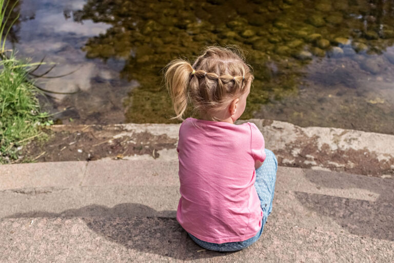 Kleines Mädchen sitzt allein am Ufer eines Teichs